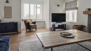 Una sala de estar con un sofá, una mesa de centro con una bandeja, un televisor sobre un mueble, una silla, un aparador y una ventana con persianas.
