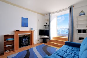 Un salón con chimenea, televisor, sofá y vista al mar a través de amplios ventanales.