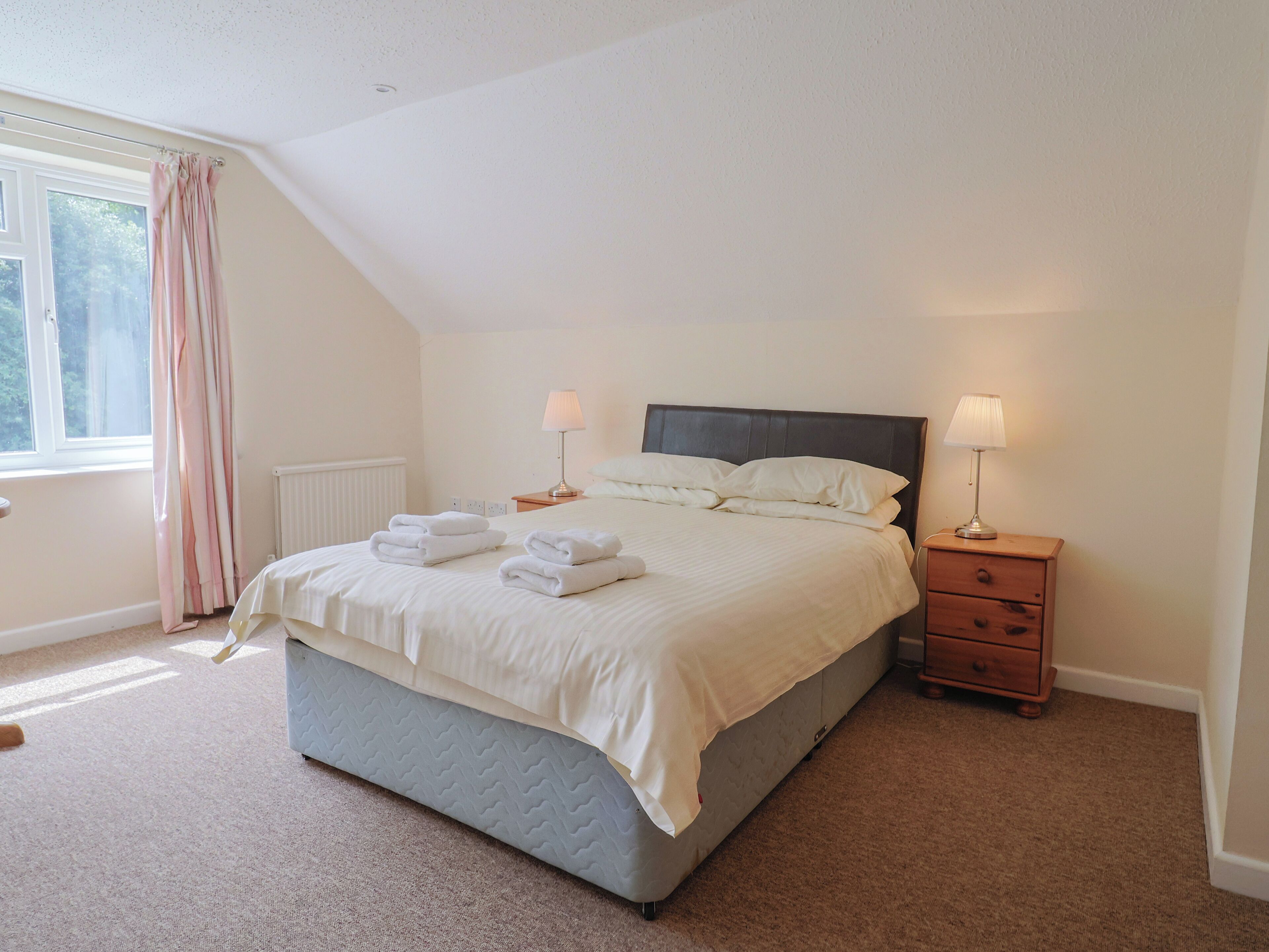A neatly made bed with folded towels, two bedside lamps, a small wooden nightstand, and a window with curtains.