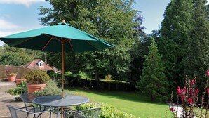 A patio with a round metal table and chairs, a green umbrella, and a garden with various plants and flowers. - Strathaven (Lyme Regis)