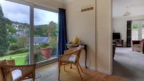 A spacious living room with a large sliding glass door leading to a garden. There is a wicker chair, a small table with a vase, and a sofa with a television in the background. - Strathaven (Lyme Regis)