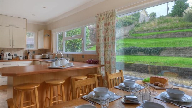 A modern kitchen with a central island, wooden stools, and a dining table set for four. - Strathaven (Lyme Regis)