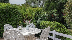 A round outdoor table with intricate metal chairs, a bottle of wine, and glasses on it, surrounded by lush greenery and a white fence.