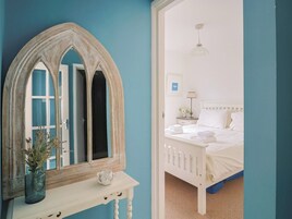 Un dormitorio con una cama, una mesita de noche con un florero, un espejo y una puerta que da a otra habitación.