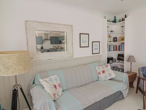 A cozy room with a sofa, a lamp, a bookshelf, and a mirror reflecting a kitchen area. - 1 Bay View Court (Lyme Regis)