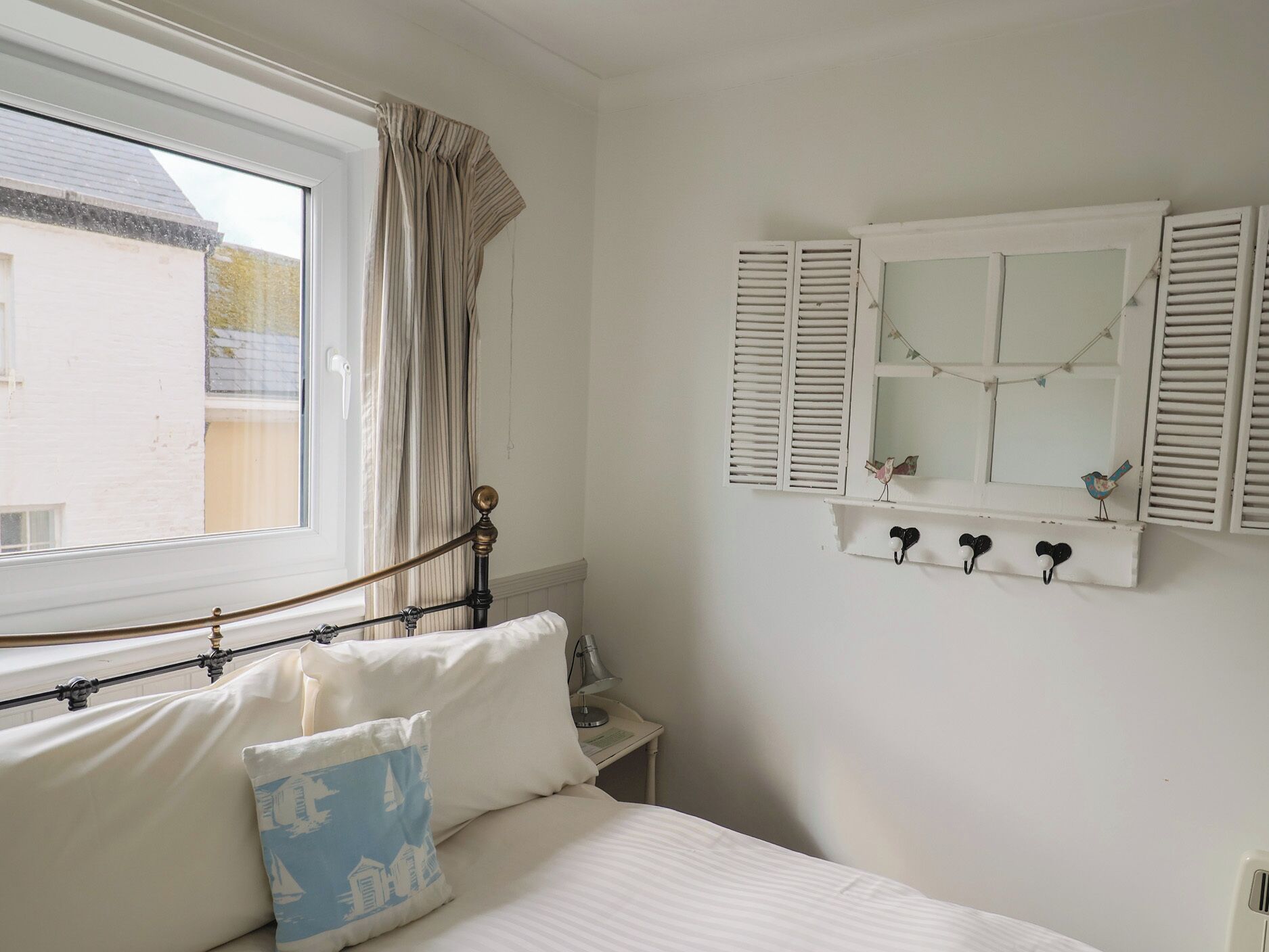 A bedroom with a bed, a window with curtains, a bedside table, a mirror with shutters, and wall-mounted hooks.