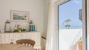 Une pièce comprenant une table en bois, une chaise, un meuble blanc orné d’objets décoratifs, un tableau encadré et une vue sur la mer, visible à travers une porte-fenêtre coulissante.