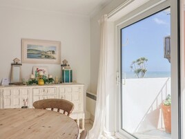 Una habitación con una mesa de madera, una silla, un armario blanco con objetos decorativos, un cuadro enmarcado y vista al mar a través de una puerta corrediza de vidrio.
