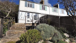 A two-story house with a stone staircase leading to the entrance, surrounded by a wooden fence and various shrubs.