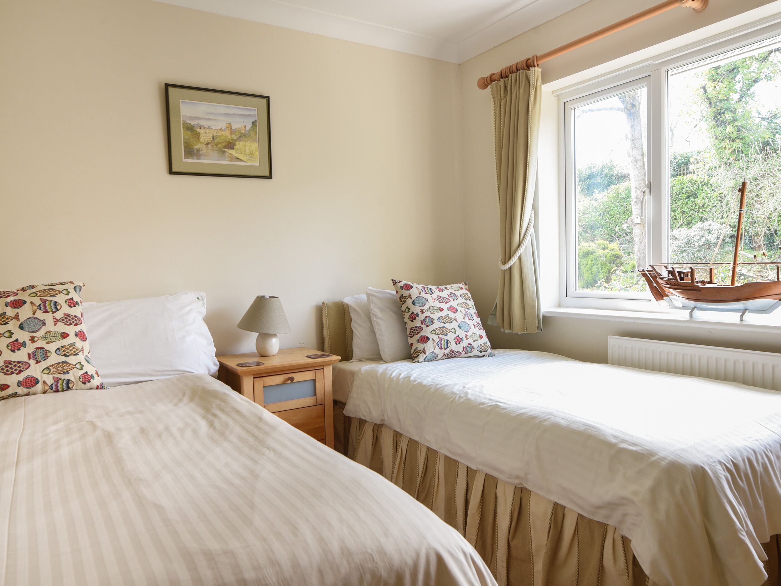 A bedroom with two beds, a nightstand with a lamp, a framed picture on the wall, and a window with a view of greenery and a boat.