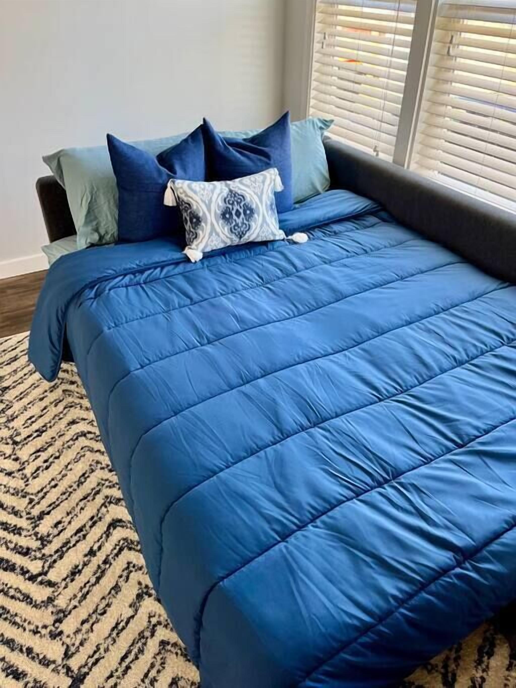 Un lit avec une couette bleue et des oreillers, un tapis à motifs et une fenêtre avec des stores.