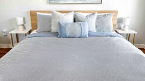 Una cama bien tendida con colcha gris y blanca estampada, flanqueada por dos mesitas de noche con lámparas blancas, y una obra enmarcada que representa un mar y un cielo tranquilos sobre la cama.