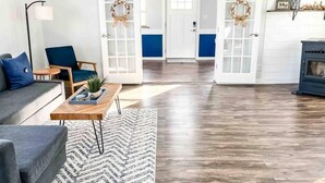 Una sala moderna con un sofá gris, un sillón azul, una mesa de centro de madera con una planta y una alfombra estampada. Hay dos puertas blancas con detalles azules y coronas decorativas colgando.