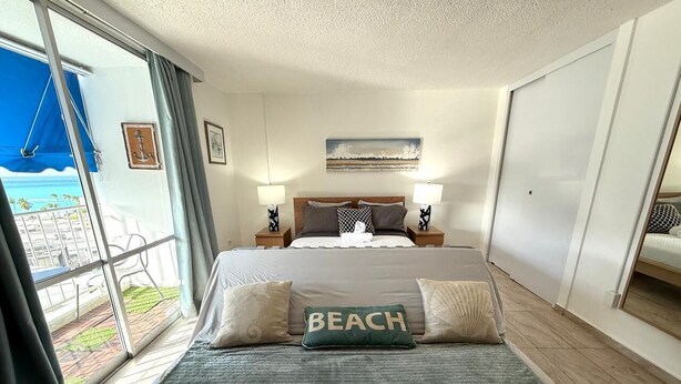 Habitación de hotel con una cama grande, dos mesitas de noche, vista a la playa y una ventana con cortinas.