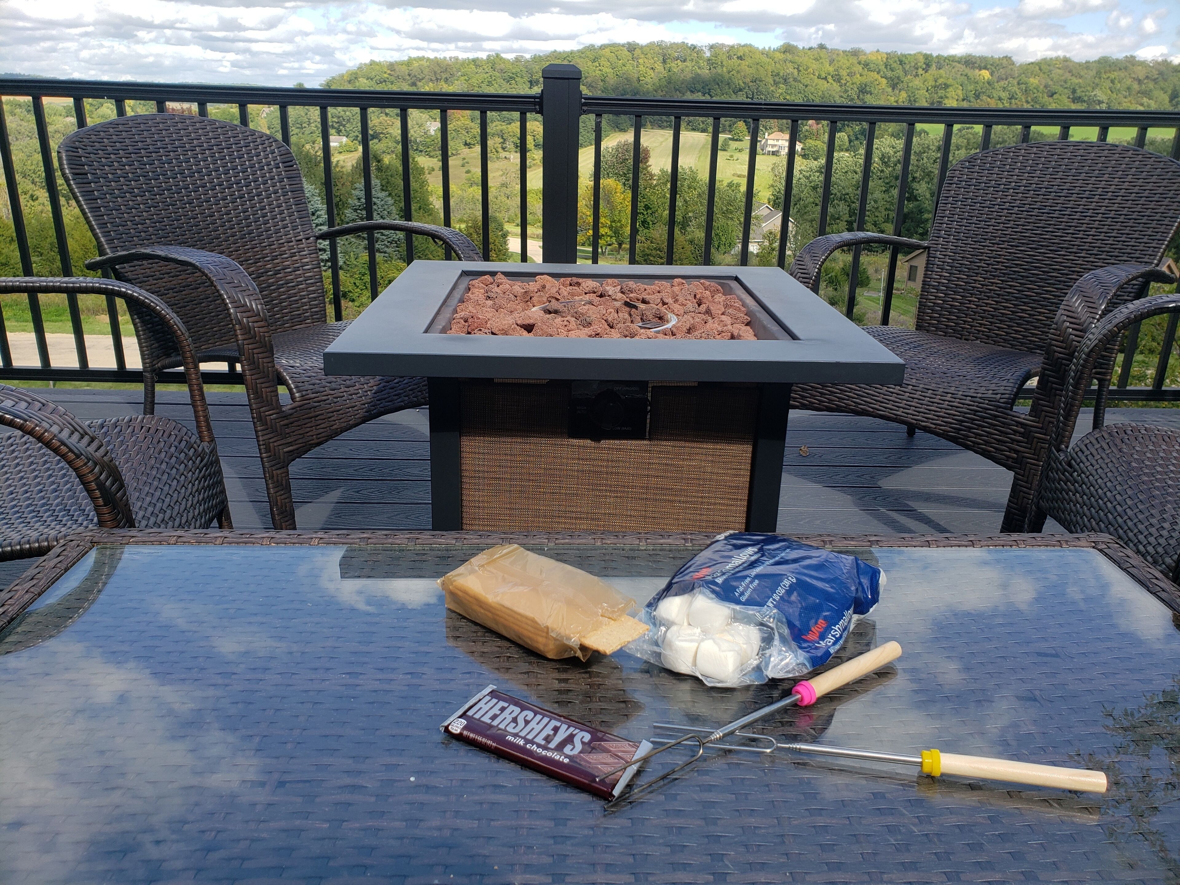A patio with wicker chairs, a glass table, a fire pit, and a bag of marshmallows.