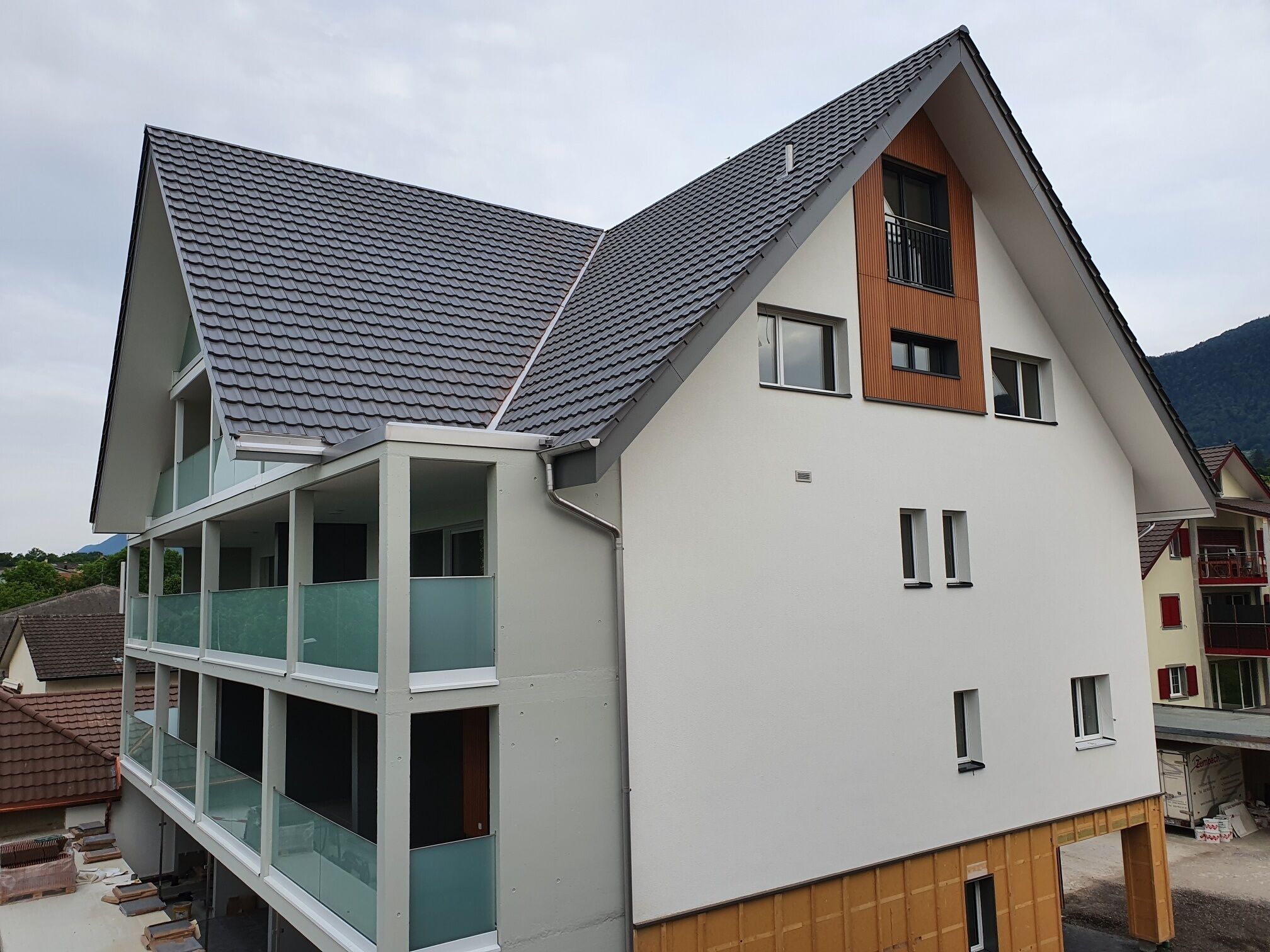 Une maison moderne à deux étages, dotée d’un toit en pente, d’un balcon au rez-de-chaussée et d’une petite terrasse en bois.