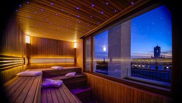 A wooden sauna with benches and a large window offering a view of a tower. - La Recova Hotel & Spa (La Punta)