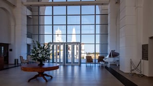 A spacious lobby with a large glass wall offering a view of a church and a bell tower. There is a round wooden table with a floral arrangement in the center, and a piano on the right. - La Recova Hotel & Spa (La Punta)