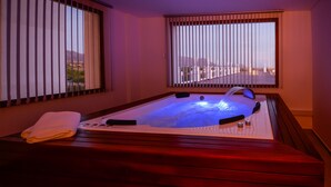 A hot tub with a wooden bench and a towel, set against a backdrop of a cityscape through large windows. - La Recova Hotel & Spa (La Punta)