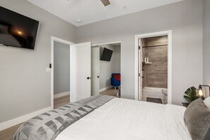 A bedroom with a bed, a TV mounted on the wall, a bathroom with a bathtub, and a chair in the corner.