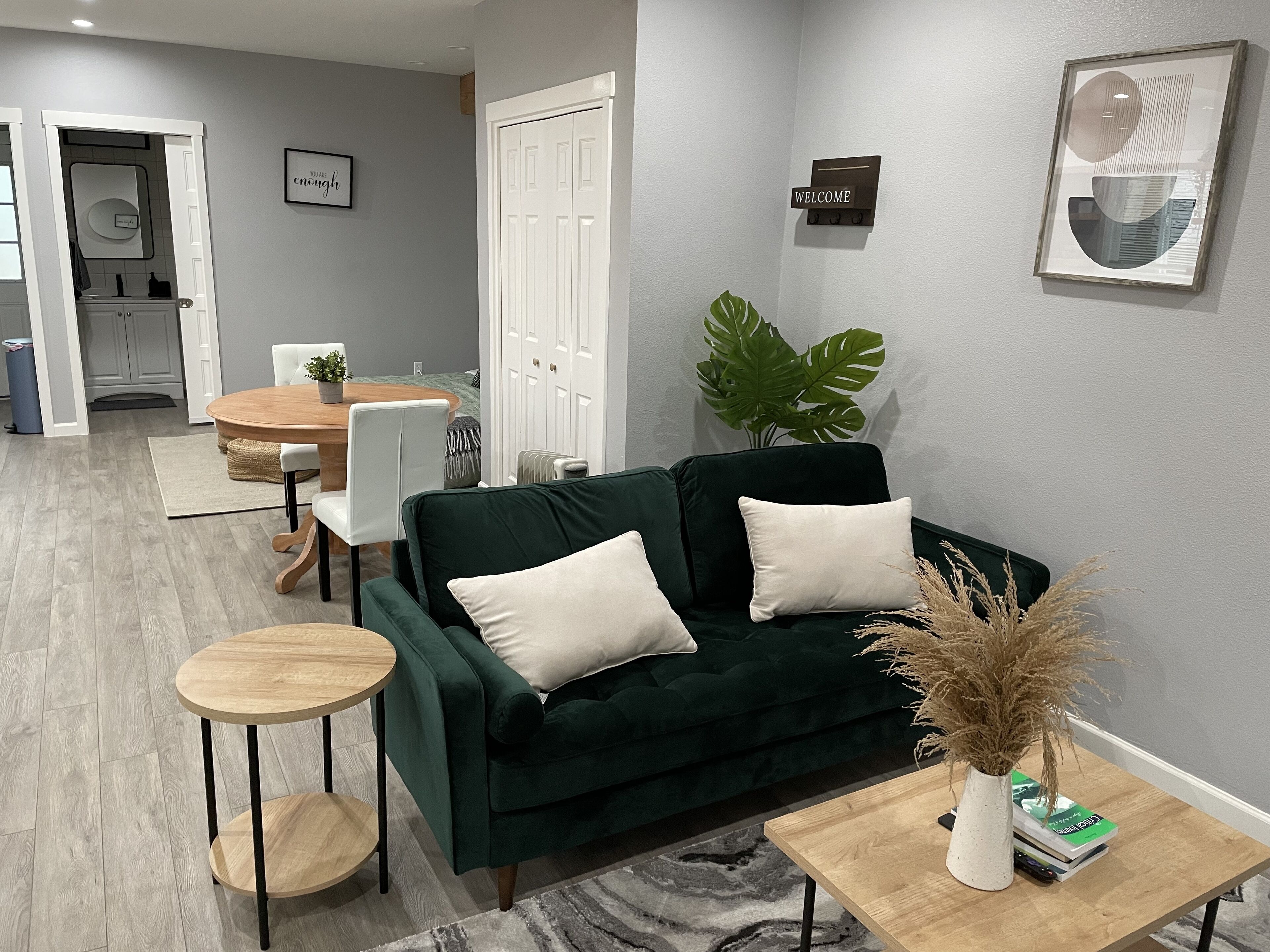 Un salon moderne avec un canapé vert foncé, une petite table d’appoint ronde en bois et une table basse. On aperçoit à l’arrière-plan un coin repas avec une table et des chaises en bois.