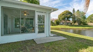 Un patio couvert avec une porte-fenêtre coulissante, une marche en béton menant à la porte, et une vue sur un étang et des arbres en arrière-plan.