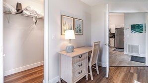 Une pièce comprenant un bureau, une chaise et une lampe. On y trouve un placard avec des vêtements suspendus et une étagère avec des serviettes. On aperçoit une cuisine au fond.