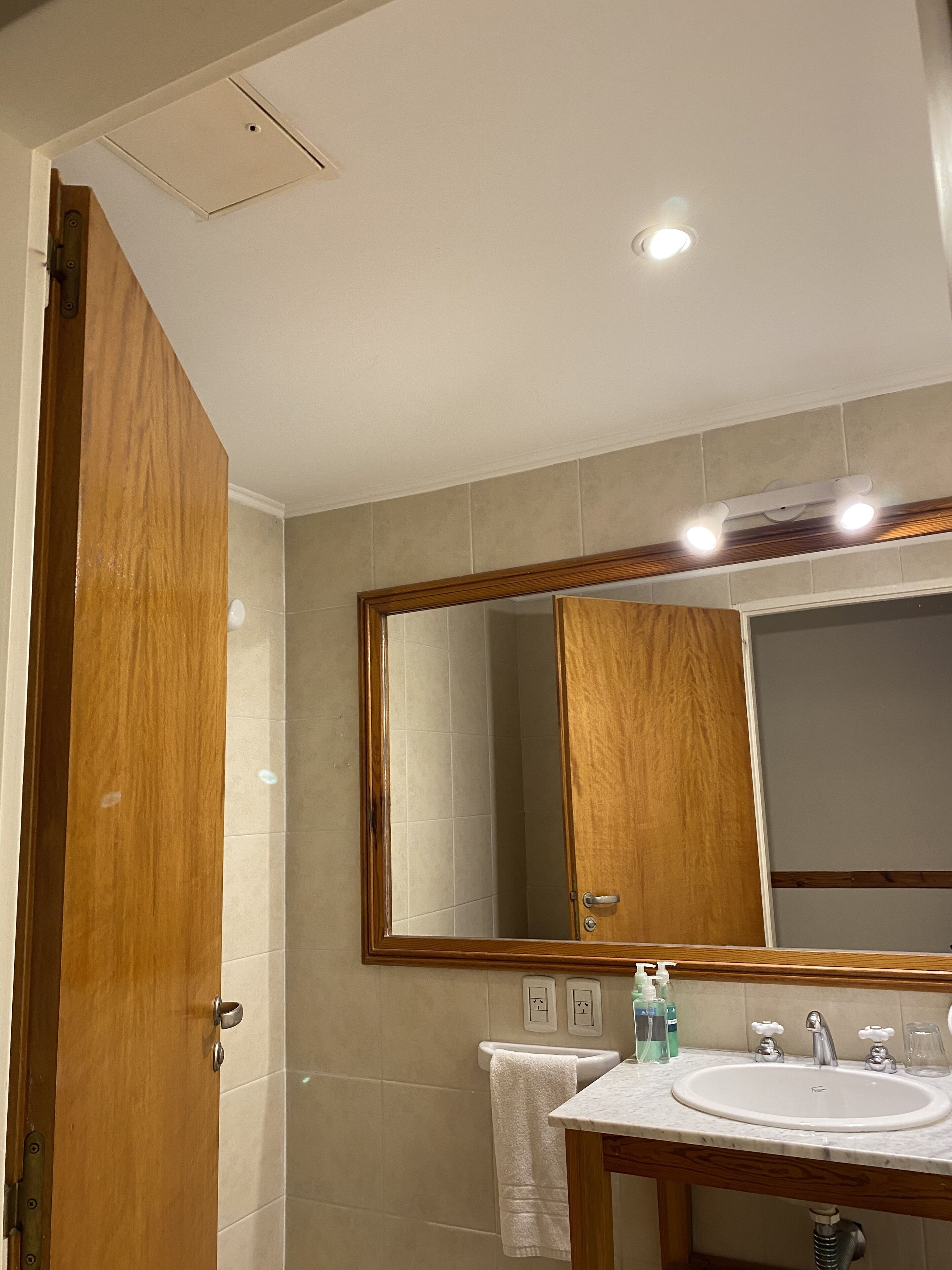 A bathroom with a wooden door, a large mirror, a sink with two faucets, and a towel hanging on the wall.