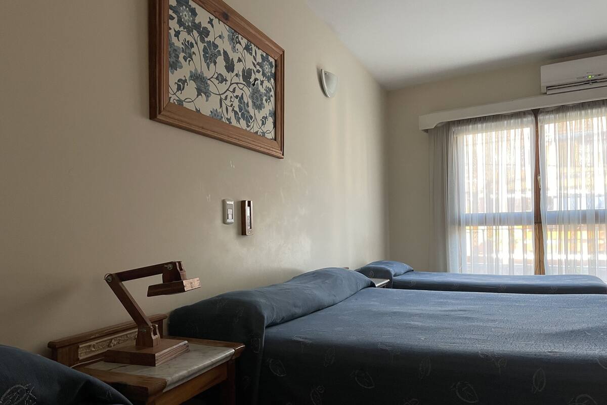 Habitación de hotel con dos camas, un escritorio con lámpara, una obra de arte floral enmarcada, una luz empotrada en la pared y una ventana con cortinas.