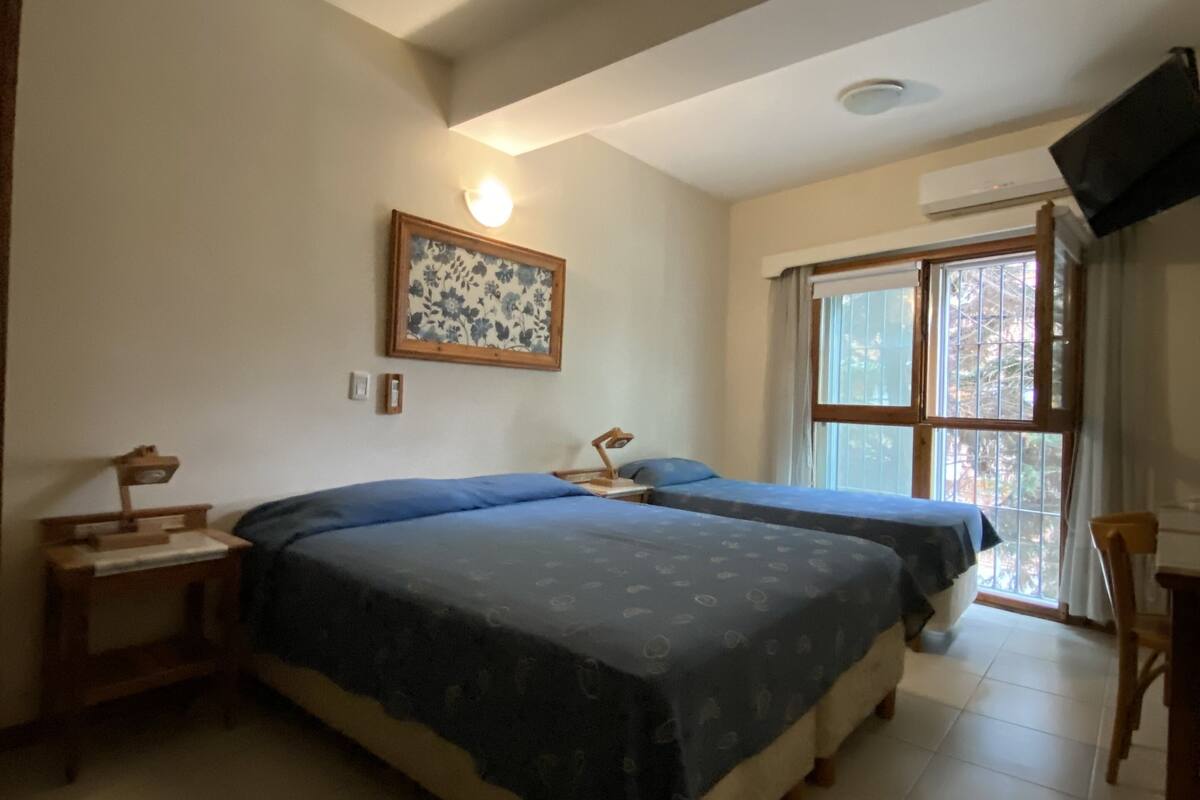 Un dormitorio con dos camas, una mesita de noche, un escritorio, una silla, un ventanal con cortinas y un televisor montado en la pared.