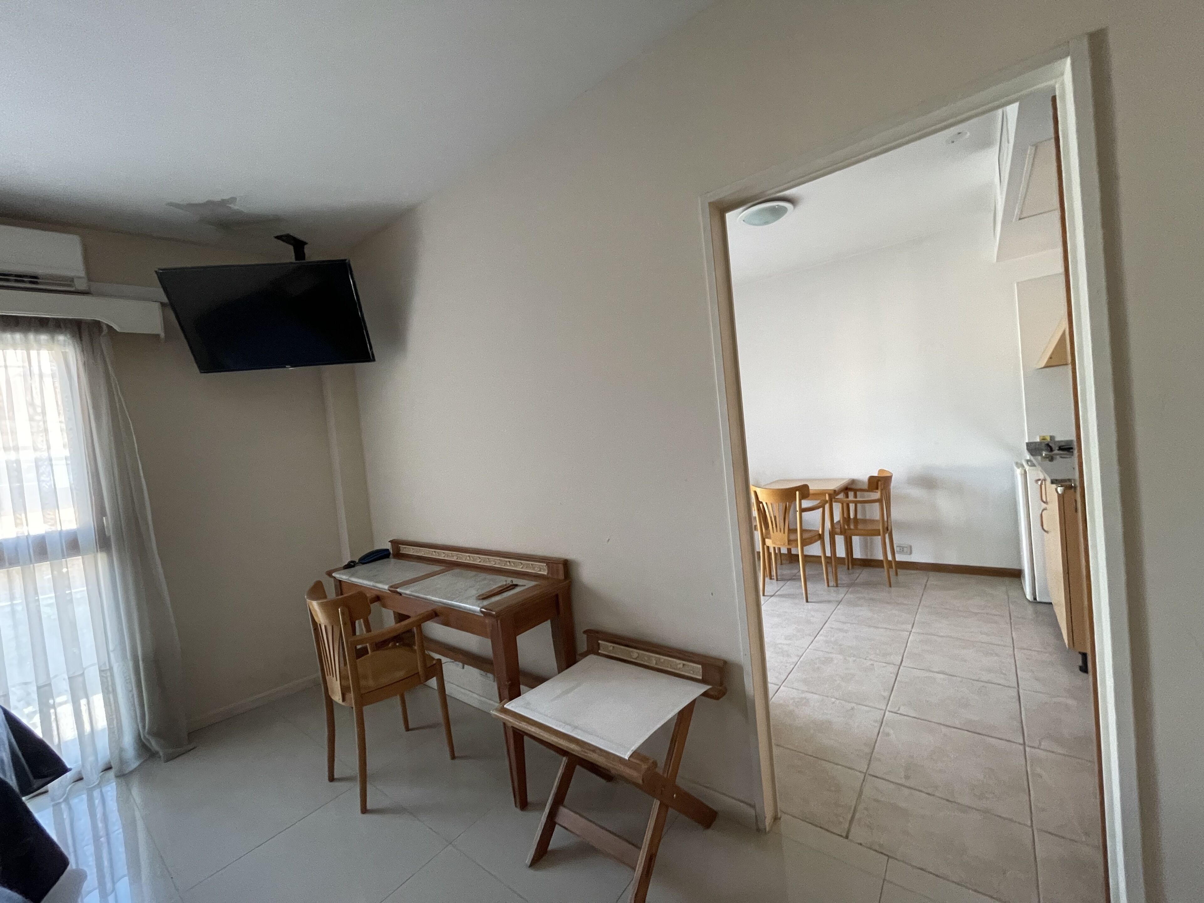 A room with a TV mounted on the wall, a dining table with chairs, and a kitchen area visible through an open doorway.