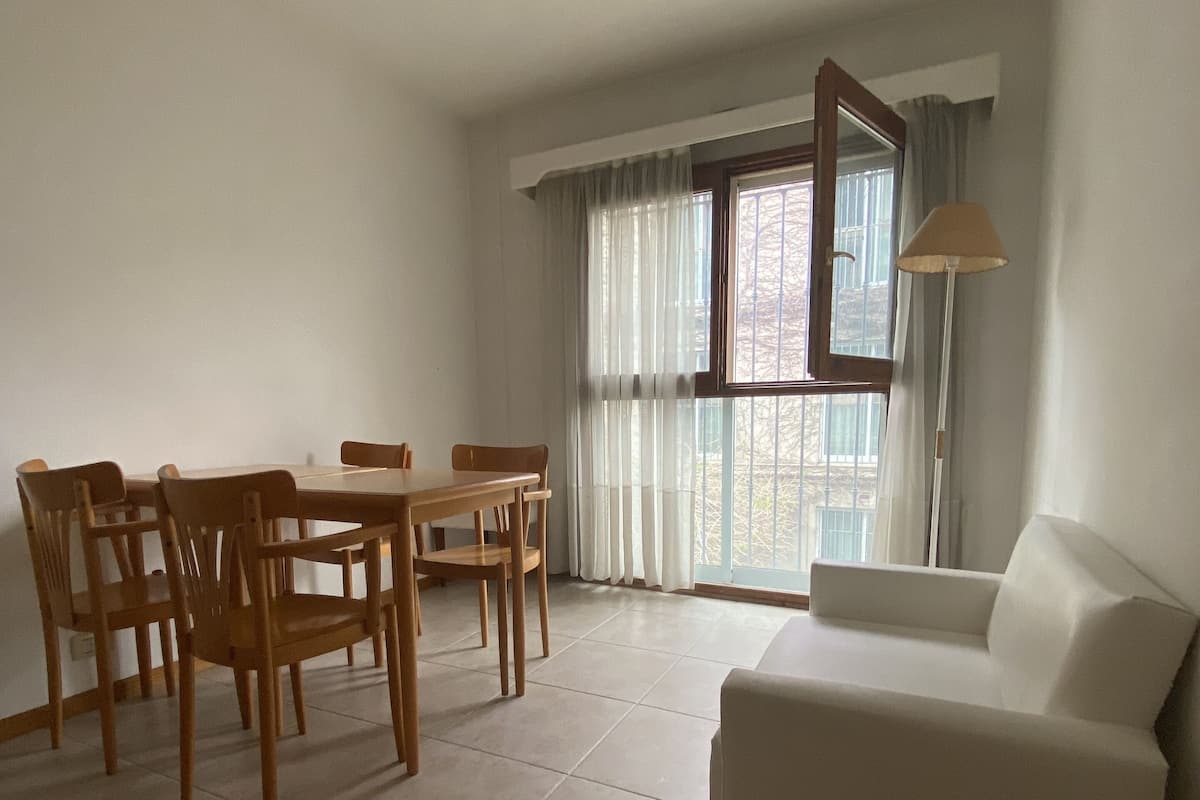 Una sala de estar con un sofá blanco, una mesa de comedor de madera y sillas. Hay una ventana con cortinas y una lámpara de pie.