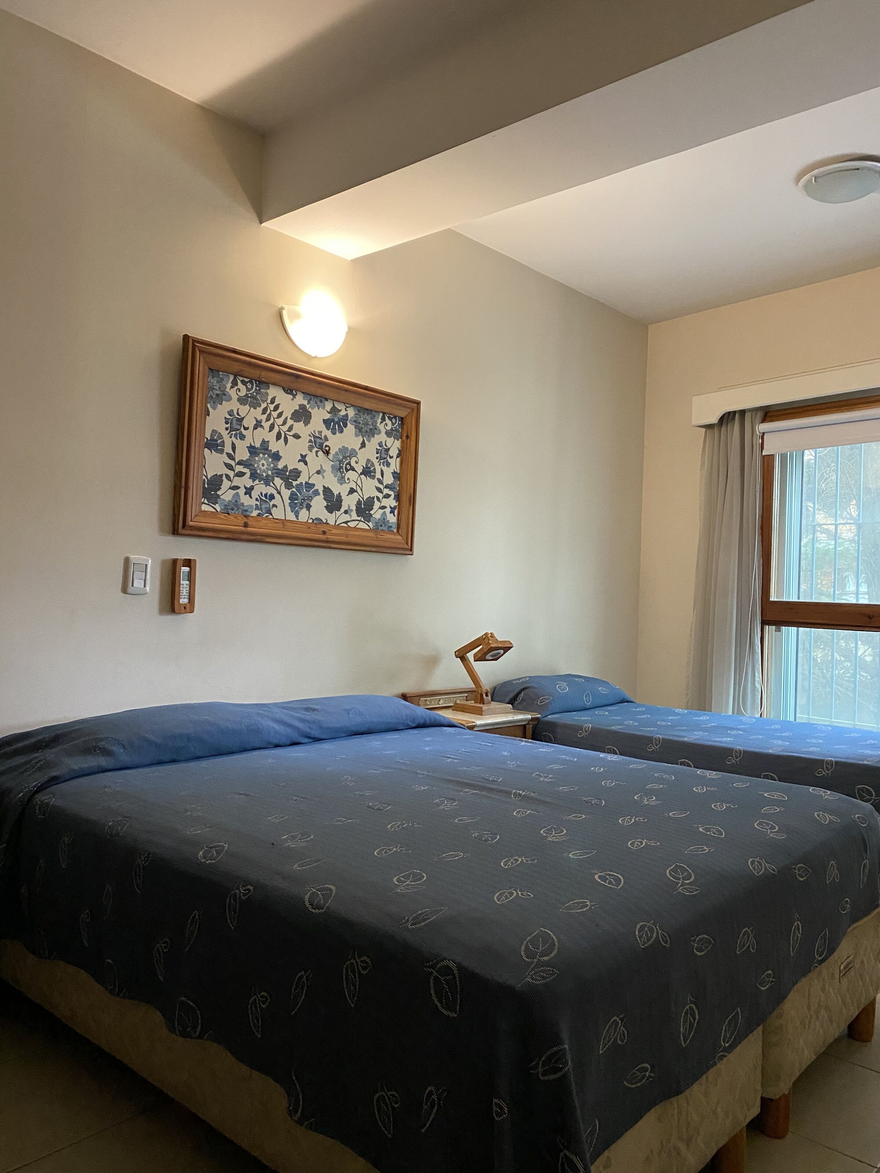 A room with two beds, a nightstand, a wall-mounted lamp, a framed floral artwork, and a window with curtains.