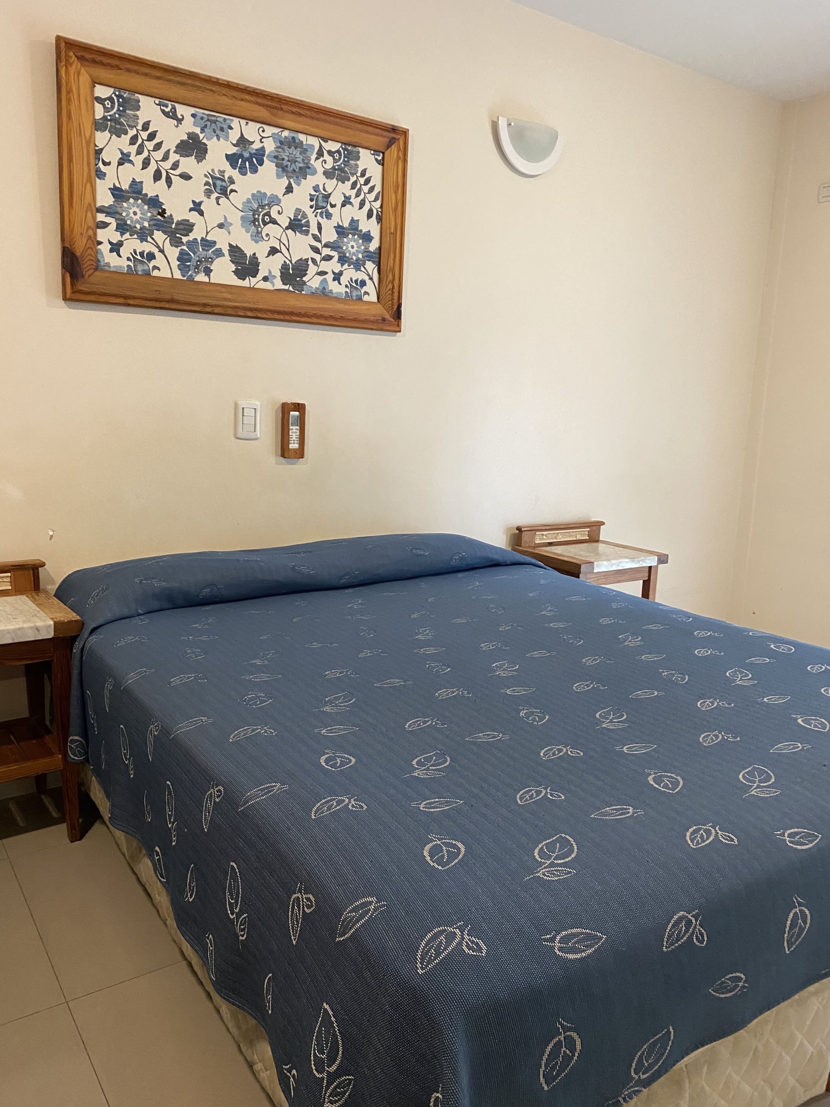 Un dormitorio con una cama, una mesita de noche, una lámpara de pared y una obra de arte floral enmarcada.