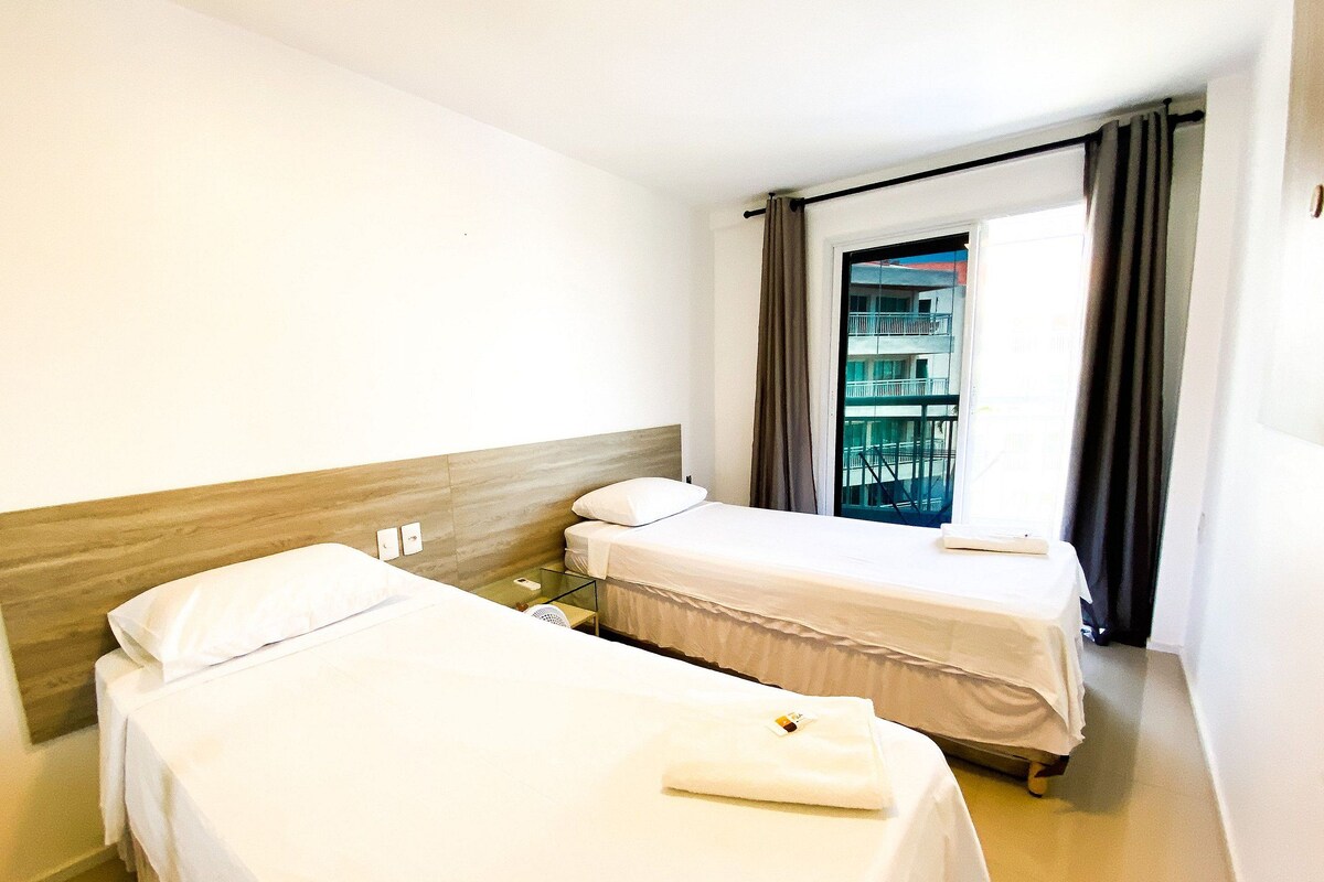 Habitación de hotel con dos camas individuales, una mesita, una ventana con cortinas y vistas a un edificio.
