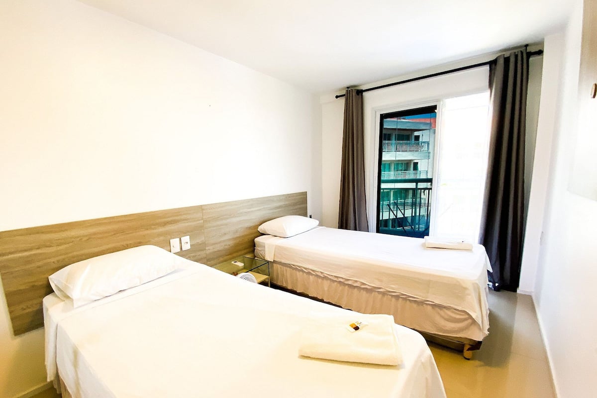 A hotel room with two single beds, a small table, a window with curtains, and a view of a building outside.