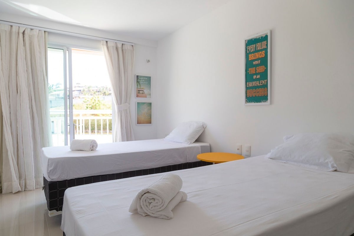 Quarto de hotel com duas camas, uma mesinha laranja e uma varanda com cortinas.