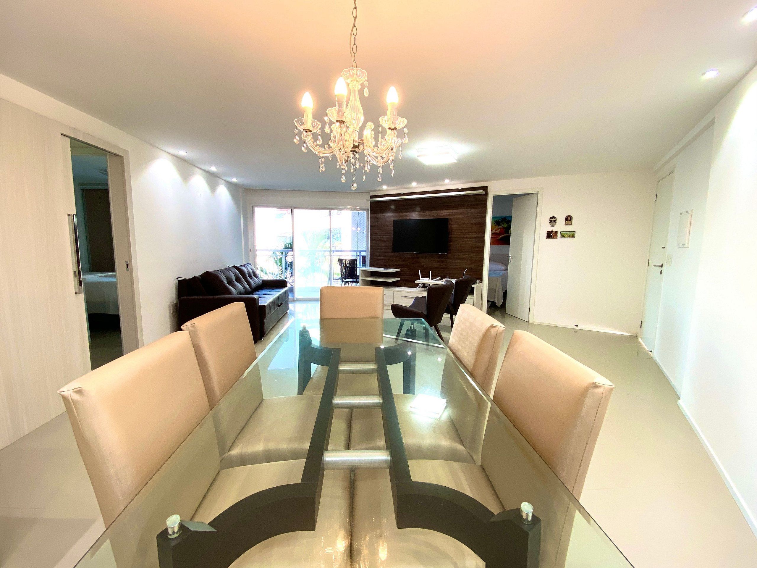 A modern living room with a glass dining table, leather sofas, a chandelier, and a TV mounted on the wall.