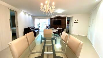A modern living room with a glass dining table, leather sofas, a chandelier, and a TV mounted on the wall.