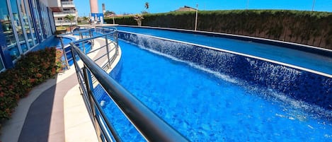 Piscina com design curvo, cercada por um edifício com varandas e céu claro.