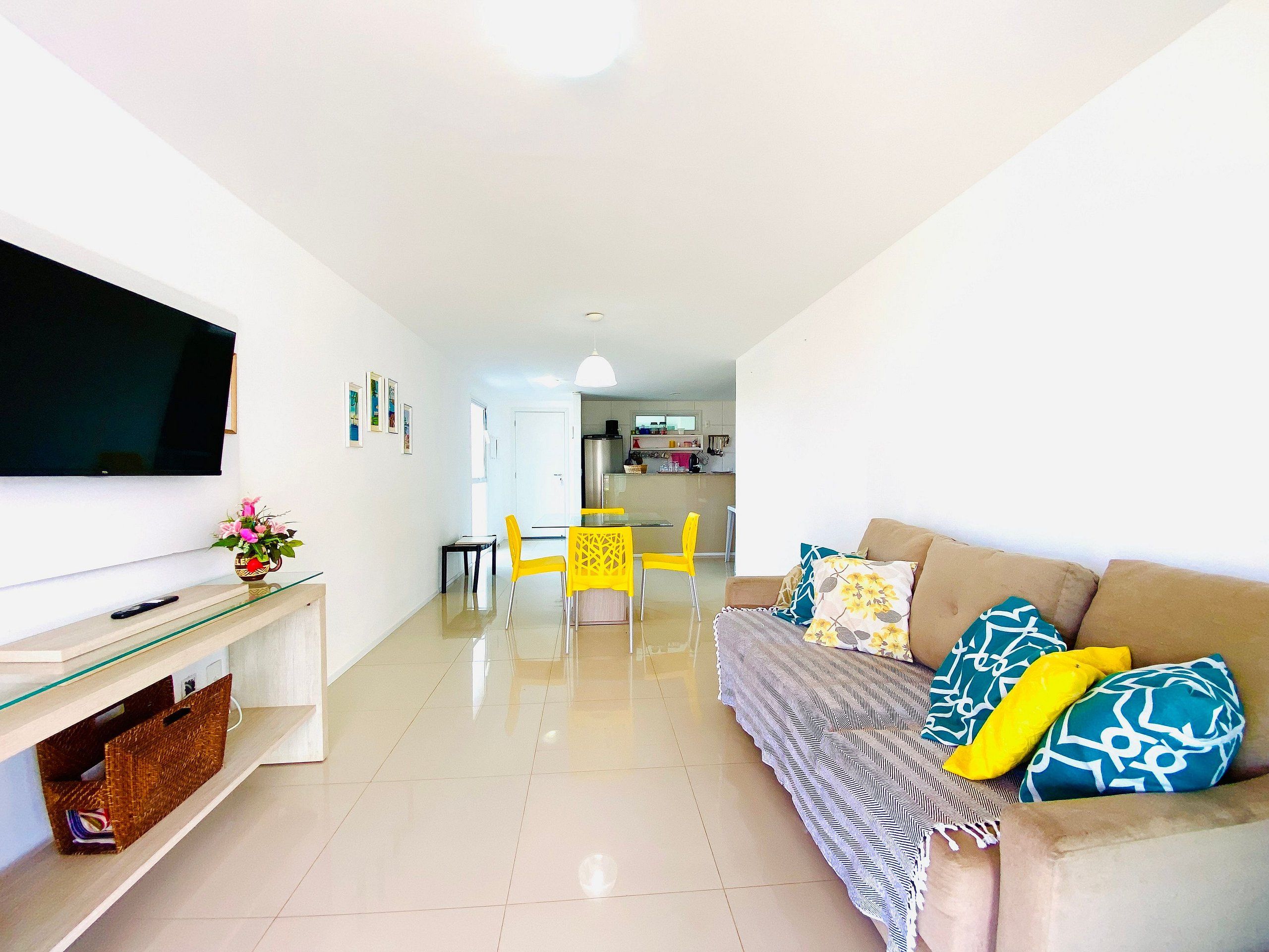 Uma sala de estar moderna com uma TV de tela plana, um sofá com almofadas estampadas, uma área de refeições com cadeiras amarelas e uma cozinha ao fundo.