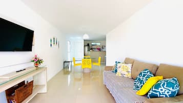 Uma sala de estar moderna com uma TV de tela plana, um sofá com almofadas estampadas, uma área de refeições com cadeiras amarelas e uma cozinha ao fundo.