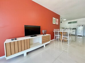 A modern living room with a flat-screen TV, wooden entertainment unit, white chairs, and a kitchen in the background.