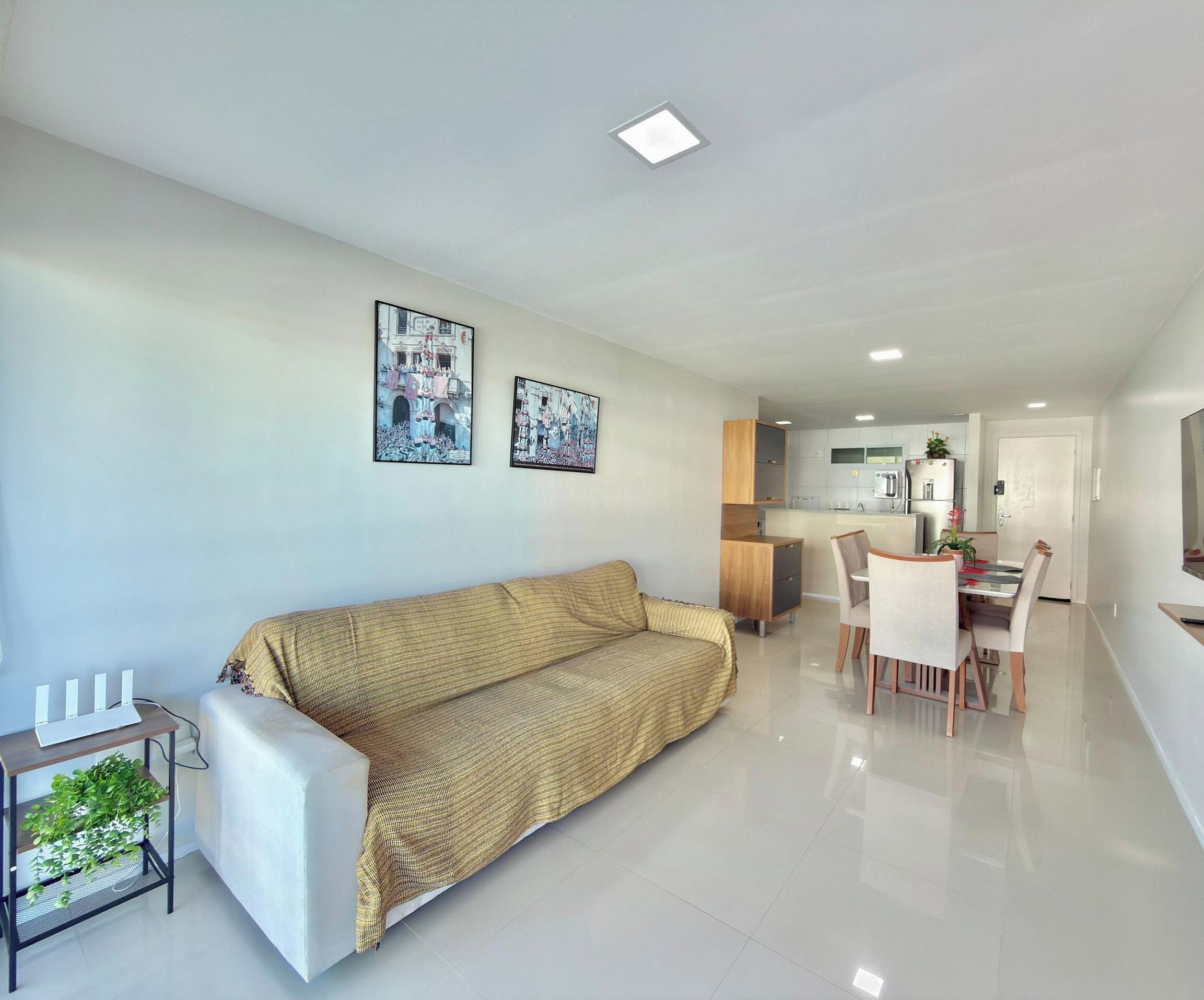 Uma sala de estar moderna com um sofá, uma área de refeições com cadeiras e uma mesa, e uma cozinha ao fundo.