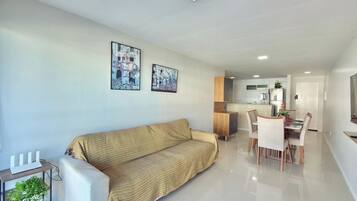 Uma sala de estar moderna com um sofá, uma área de refeições com cadeiras e uma mesa, e uma cozinha ao fundo.