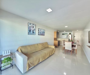 A modern living room with a sofa, a dining area with chairs and a table, and a kitchen in the background. - Solarium Residence Resort (Aquiraz)