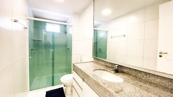 Casa de banho com cabine de chuveiro em vidro, sanita branca, lavatório com bancada em granito e um espelho grande.