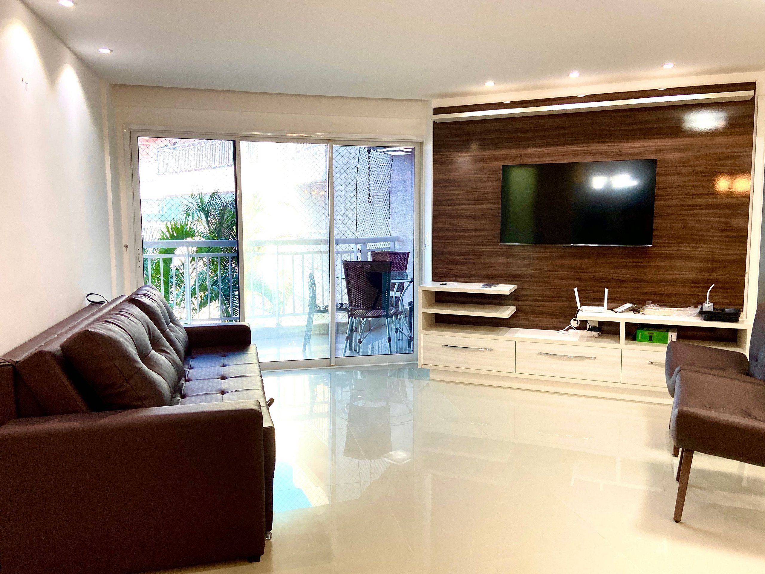 Una sala moderna con sofá, televisión y un balcón con mobiliario exterior.