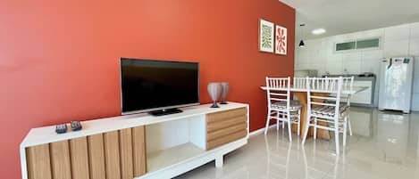 A modern living room with a flat-screen TV, wooden entertainment unit, white chairs, and a kitchen in the background.