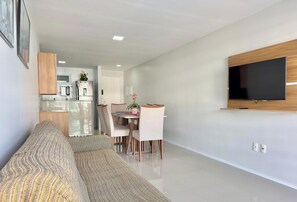 Una sala de estar moderna con sofá, mesa de comedor, sillas, televisor montado en la pared y una cocina al fondo.